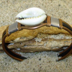 Könnte beinhalten: Ein Kupfer- und Silberarmband mit einem schwarzen Lederband und einem weißen Kaurischalen-Akzent.