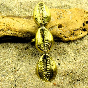 May include: A gold-colored ring featuring three vertically aligned, stylized seashell shapes. The ring is set against a sandy background with a piece of weathered, tan-colored rock. The ring's design evokes a coastal or nautical theme.