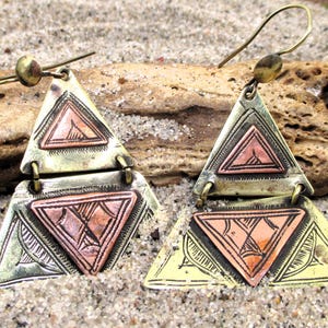 May include: A pair of brass-toned earrings with a geometric design. Each earring features two connected triangles, with a copper-colored triangle in the center. The earrings have a hook closure and are displayed on a sandy surface.