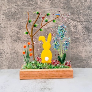 以下が含まれることがあります： 青い鳥、オレンジの花、紫の花が咲く庭園のシーンの中に、黄色いガラス製のウサギが立っています。シーンは透明なガラスパネルに囲まれ、木製の台座に載せられています。