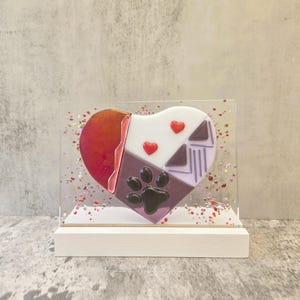 May include: A heart-shaped decorative piece with a paw print design. The heart is divided into sections of red, white, purple, and lavender, with two red heart accents. The piece is encased in clear acrylic and sits on a white wooden base.