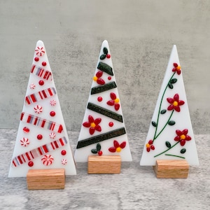 Puede incluir: Tres árboles de Navidad decorativos de cristal con fondos blancos y bases de madera. Un árbol presenta rayas rojas y blancas de bastón de caramelo, otro tiene flores rojas y rayas verdes, y el tercero tiene flores rojas sobre tallos verdes.