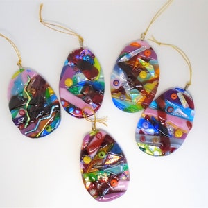 May include: Five colorful glass ornaments shaped like eggs. Each ornament is decorated with a variety of colors and textures, including red, blue, green, yellow, and purple. The ornaments are strung with gold cords.