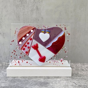 Può includere: Un'opera d'arte decorativa in vetro a forma di cuore. Il cuore è diviso in sezioni bordeaux, bianche e rosse, con una freccia rossa e un cuore bianco. Due uccelli blu si posano sopra un cuore bianco. Il pezzo è racchiuso in una custodia di acrilico trasparente su una base bianca.