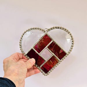 Pode incluir: Um ornamento de vidro colorido em forma de coração com um design geométrico que apresenta peças de vidro vermelho e transparente. O coração é contornado com contas prateadas.