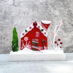 May include: A festive Christmas decoration featuring a red gingerbread house with white accents, candy canes, and a green tree. The house is set against a snowy backdrop, mounted on a white wooden base, creating a charming holiday scene.
