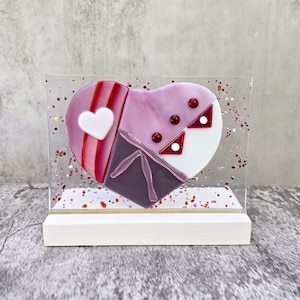 Può includere: Un cuore di vetro con un design a mosaico rosa, viola e bianco. Il cuore è circondato da glitter rosso e bianco. Il cuore è su un supporto bianco.