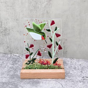 May include: A stained glass hummingbird with red and green accents is perched on a branch with red flowers. The glass piece is mounted on a wooden stand.