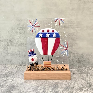 May include: A patriotic glass art piece featuring a red, white, and blue hot air balloon design with stars and stripes. The artwork includes a small Uncle Sam figure and fireworks, all set on a wooden base. The piece is ideal for Independence Day.