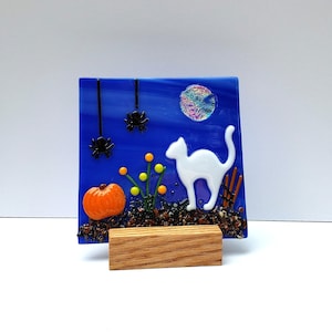 May include: A blue glass tile with a white cat, a pumpkin, and spiders hanging from the ceiling. The tile has a glittery texture and is set on a wooden stand.