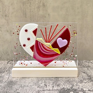 Può includere: Una scultura di cuore in vetro con pezzi di vetro rosso, bianco e rosa. Il cuore è circondato da piccole paillettes colorate. La scultura è su una base di legno bianco.
