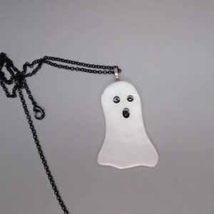 Puede incluir: Collar con colgante de fantasma blanco y cadena negra. El fantasma tiene tres ojos de cuentas negras y una boca abierta negra. El colgante es nacarado y tiene un ligero tono rosado.