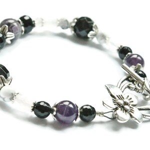 May include: A black, white, and purple beaded bracelet with a silver butterfly clasp. The bracelet features amethyst, black onyx, and white howlite beads.