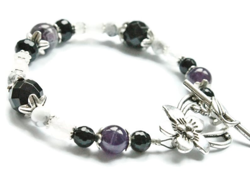 May include: A black, white, and purple beaded bracelet with a silver butterfly clasp. The bracelet features amethyst, black onyx, and white howlite beads.