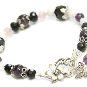 May include: A bracelet with amethyst, black onyx, and white beads. The bracelet has a silver clasp with a flower and butterfly design.