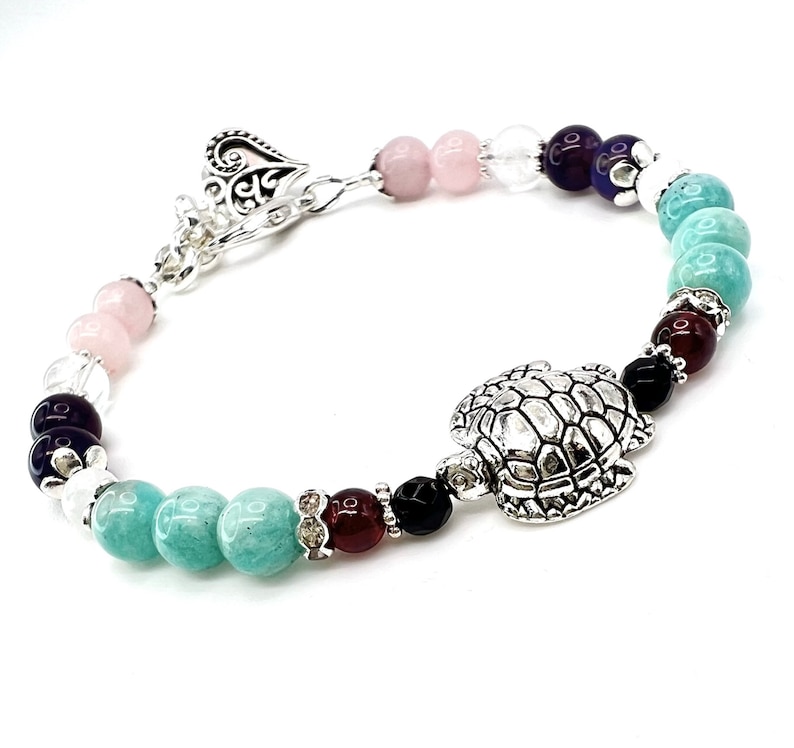 May include: A beaded bracelet featuring a silver turtle charm and a heart-shaped clasp. The bracelet incorporates a variety of colorful beads, including pink, clear, purple, turquoise, black, and burgundy. The beads are strung together with a silver chain.
