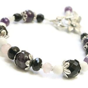 May include: A bracelet with black, white, and purple beads. The beads are interspersed with silver flower charms. The bracelet is made of natural stones and is designed to be worn on the wrist.