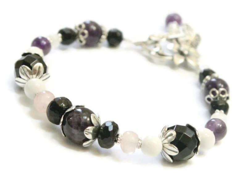 May include: A bracelet with black, white, and purple beads. The beads are interspersed with silver flower charms. The bracelet is made of natural stones and is designed to be worn on the wrist.