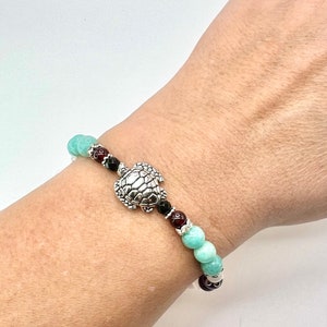 May include: A beaded bracelet featuring a silver turtle charm. The bracelet includes turquoise, black, and red beads. The bracelet is worn on a wrist.