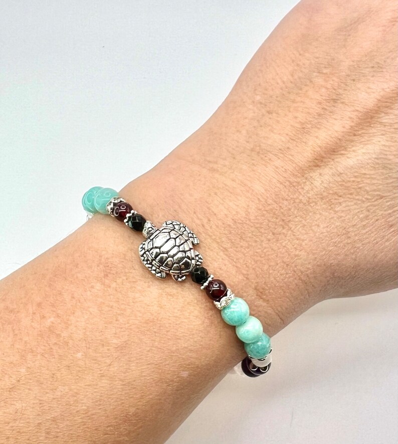 May include: A beaded bracelet featuring a silver turtle charm. The bracelet includes turquoise, black, and red beads. The bracelet is worn on a wrist.