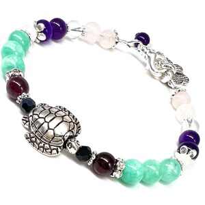 May include: A beaded bracelet featuring a silver turtle charm and a variety of colorful gemstones. The bracelet includes green, purple, clear, and pink beads, with silver accents. The bracelet is approximately 7 inches in length.