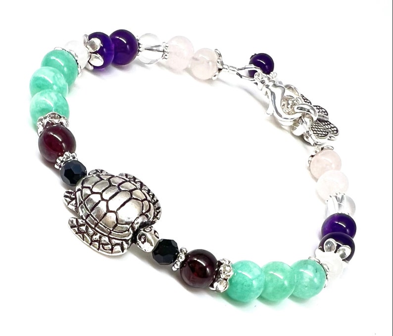 May include: A beaded bracelet featuring a silver turtle charm and a variety of colorful gemstones. The bracelet includes green, purple, clear, and pink beads, with silver accents. The bracelet is approximately 7 inches in length.