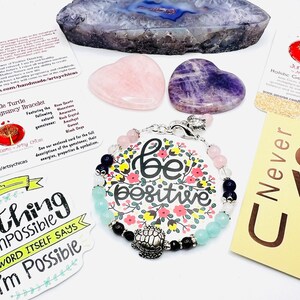 May include: A collection of gemstone jewelry and crystals. A bracelet with pink, blue, and black beads and a 'be positive' charm is featured. Also included are heart-shaped rose quartz and amethyst stones, and a sticker that says 'Nothing is impossible'.