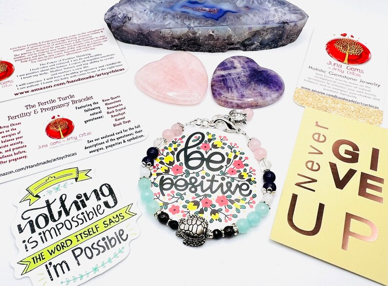 May include: A collection of gemstone jewelry and crystals. A bracelet with pink, blue, and black beads and a 'be positive' charm is featured. Also included are heart-shaped rose quartz and amethyst stones, and a sticker that says 'Nothing is impossible'.