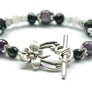 May include: A silver bracelet with a flower charm, featuring amethyst, black onyx, and white beads.