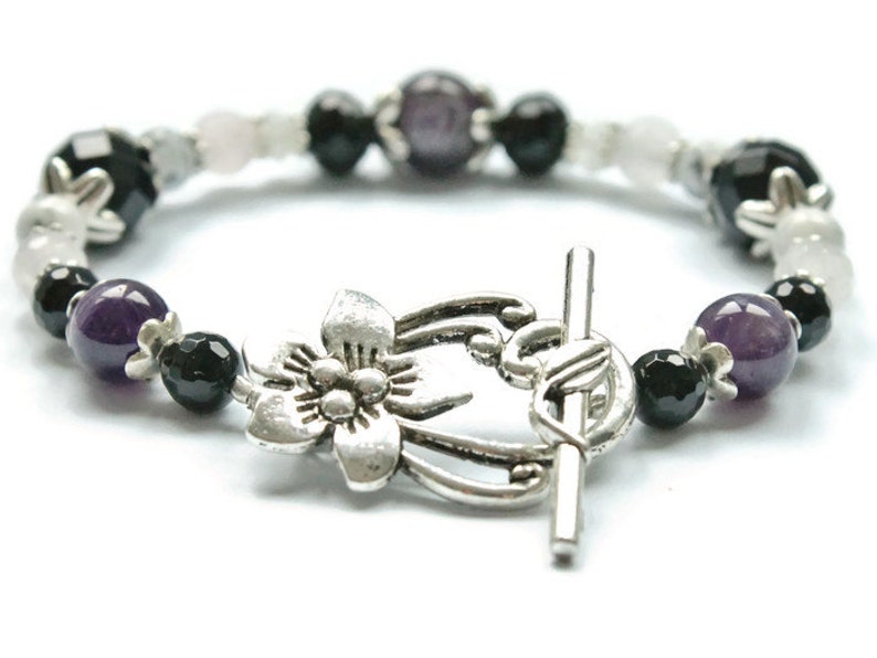 May include: A silver bracelet with a flower charm, featuring amethyst, black onyx, and white beads.