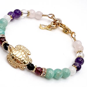 May include: A gold-colored bracelet featuring a turtle charm and a variety of colorful beads. The beads include shades of purple, pink, green, and black, with gold accents. The bracelet is designed with an adjustable clasp.