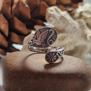 May include: A silver open-style ring featuring a hummingbird motif with copper-toned wings and engraved floral scrollwork. The band is stamped with 925, indicating sterling silver composition, and has a wrap-around design with rounded ends.