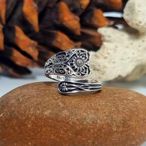 May include: A silver-coloured adjustable ring with an ornate floral design. The ring features a sunflower-like centre and intricate filigree patterns. The band is open and adjustable, resting on a brown stone.