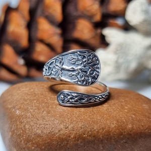 May include: A silver ring with an open design. The oval top features a detailed tree of life design. The band has a Celtic knot pattern. The ring is displayed on a brown stone. The ring is a piece of jewellery.