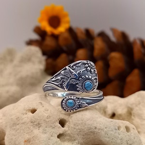May include: A silver ring with a hummingbird and sunflower design. The ring has turquoise-coloured stones and is set against a blurred background. The ring is a wrap-around style.