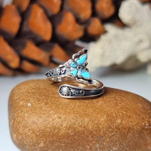 May include: A silver ring featuring a butterfly design with turquoise stone detailing. The ring sits on a brown stone. The ring has a wrap-around design with floral and sun details. The ring is a piece of jewellery.
