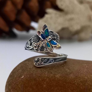 May include: A silver ring featuring a dragonfly design. The dragonfly is crafted from blue and green opal and has gold accents. The ring is displayed on a brown stone.