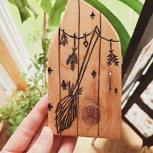 May include: A wooden door with a carved image of a broom and hanging herbs. The door is painted in a light brown color and has a rustic look.