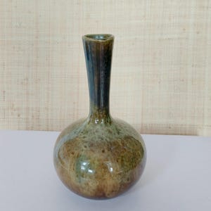 Peut inclure: Un petit vase en céramique fait à la main, avec une base arrondie et un col fin et allongé. Le vase présente une finition brillante avec un mélange de tons terreux, dont le marron, le vert et le bleu, créant une esthétique organique unique. Le vase est idéal pour les fleurs.