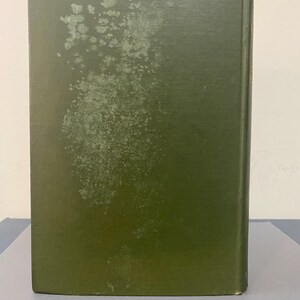 May include: A hardcover book with a faded olive green cover. The cover shows signs of wear and tear, with areas of discoloration and spots. The spine is visible, indicating the book's thickness and binding. The book is likely vintage or used.