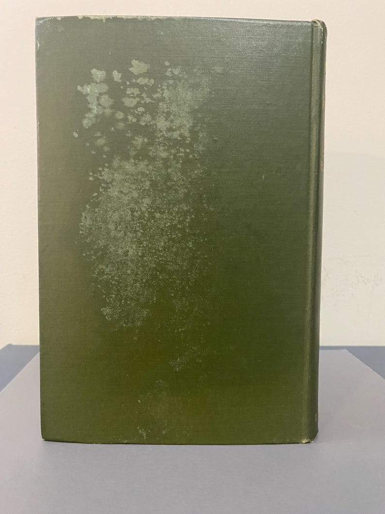 May include: A hardcover book with a faded olive green cover. The cover shows signs of wear and tear, with areas of discoloration and spots. The spine is visible, indicating the book's thickness and binding. The book is likely vintage or used.