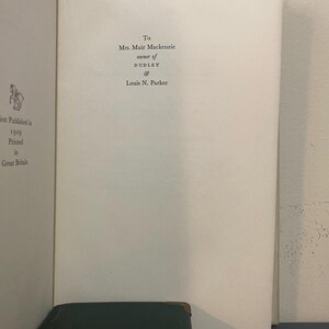 May include: Open book page with text: "To Mrs. Muir Mackenzie, owner of Dudley & Louis N. Parker." The book is open to a white page with text. The bottom of the image shows a dark green book with gold lettering.