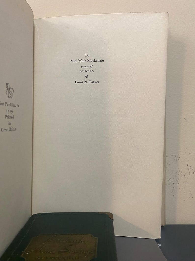 May include: Open book page with text: "To Mrs. Muir Mackenzie, owner of Dudley & Louis N. Parker." The book is open to a white page with text. The bottom of the image shows a dark green book with gold lettering.