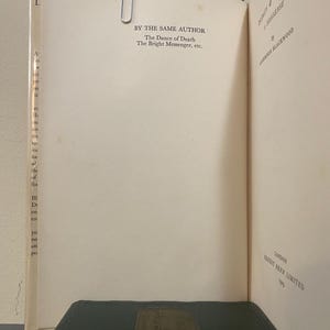 May include: Open book with text: "BY THE SAME AUTHOR The Dance of Death The Bright Messenger, etc." A metal paper clip holds the pages together. A dark green book with gold accents is at the bottom. The book is titled "HODGE & GILDEROY".