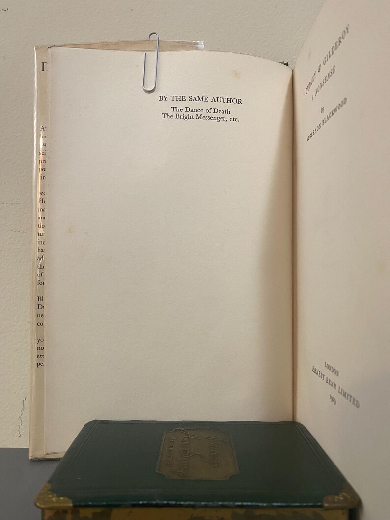 May include: Open book with text: "BY THE SAME AUTHOR The Dance of Death The Bright Messenger, etc." A metal paper clip holds the pages together. A dark green book with gold accents is at the bottom. The book is titled "HODGE & GILDEROY".