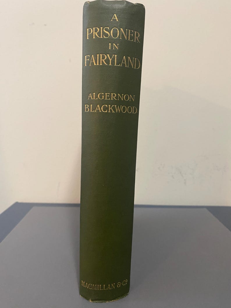 May include: A vintage hardcover book titled "A Prisoner in Fairyland" by Algernon Blackwood. The book has a dark green cover with gold lettering. The publisher's name, Macmillan & Co, is at the bottom.