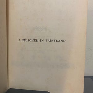 May include: Open book page with the title "A Prisoner in Fairyland" printed in black. The paper is aged with a cream color. A dark box is visible at the bottom of the image.