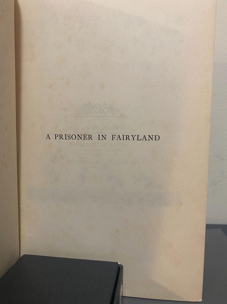 May include: Open book page with the title "A Prisoner in Fairyland" printed in black. The paper is aged with a cream color. A dark box is visible at the bottom of the image.