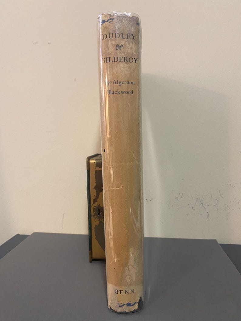 May include: A vintage book titled "Dudley & Gilderoy" by Algernon Blackwood, published by Benn. The book is wrapped in clear plastic. The cover is a light tan color with blue text.