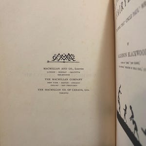 May include: Open book with the title "Prison FairyLand" by Algernon Blackwood. The book's pages are off-white with text and a decorative emblem. The Macmillan Company logo is visible.